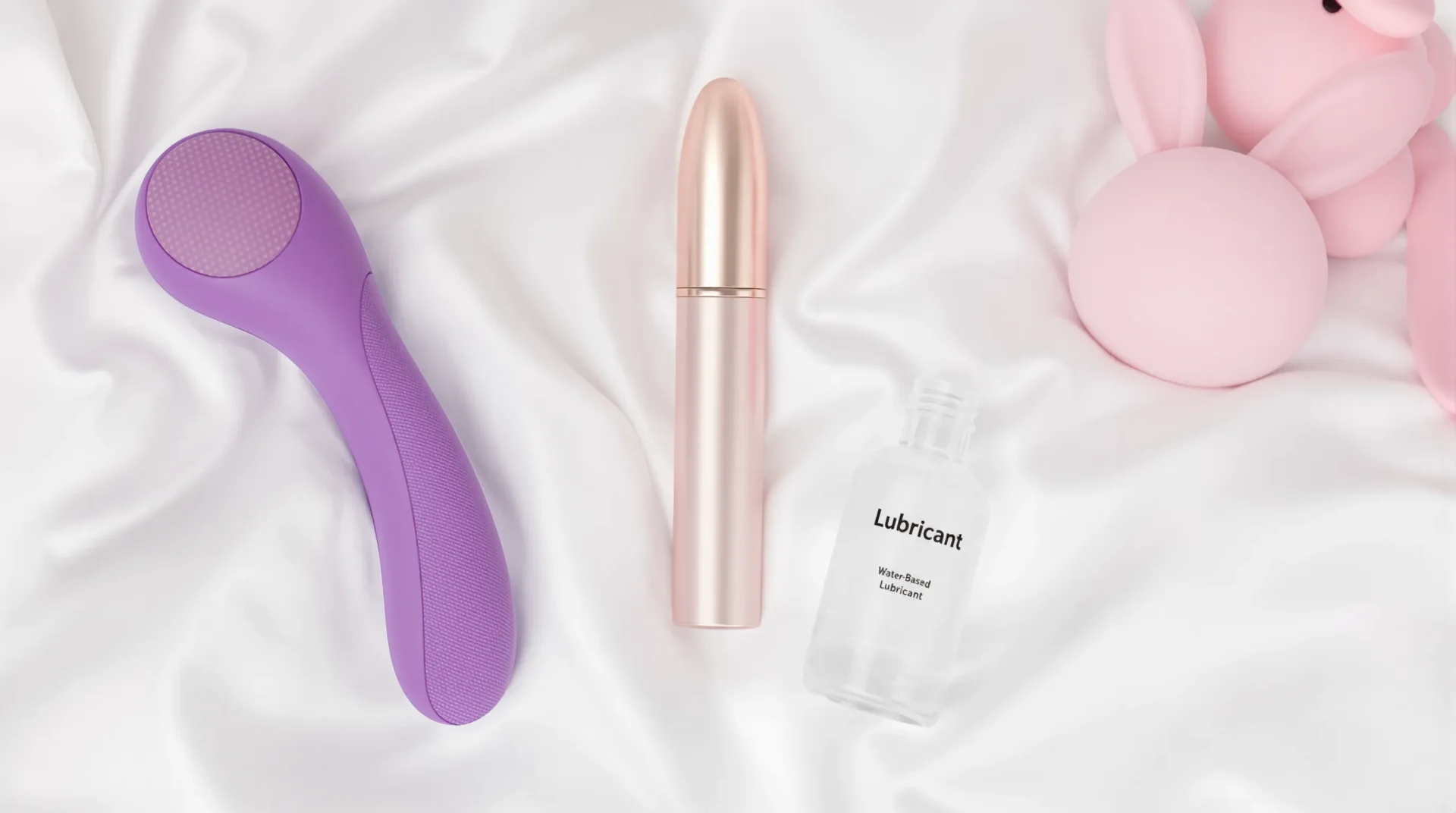 Flat-lay photo of three vibrators—a curved purple G-spot toy, a rose-gold bullet, and a pink rabbit—resting on soft linen sheets with a bottle of water-based lubricant beside them.